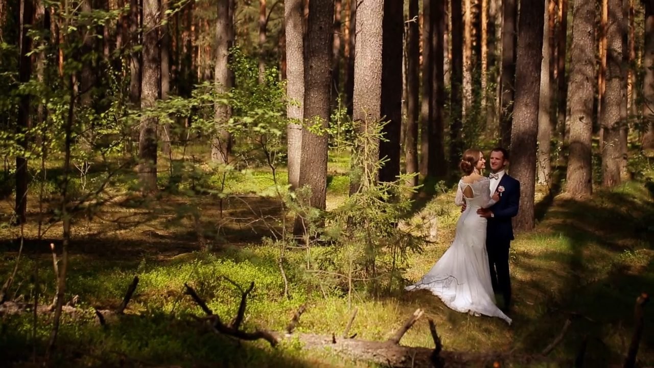 A bride and groom embrace in a sun-dappled forest. The bride wears a white wedding dress, and the groom is in a blue suit.