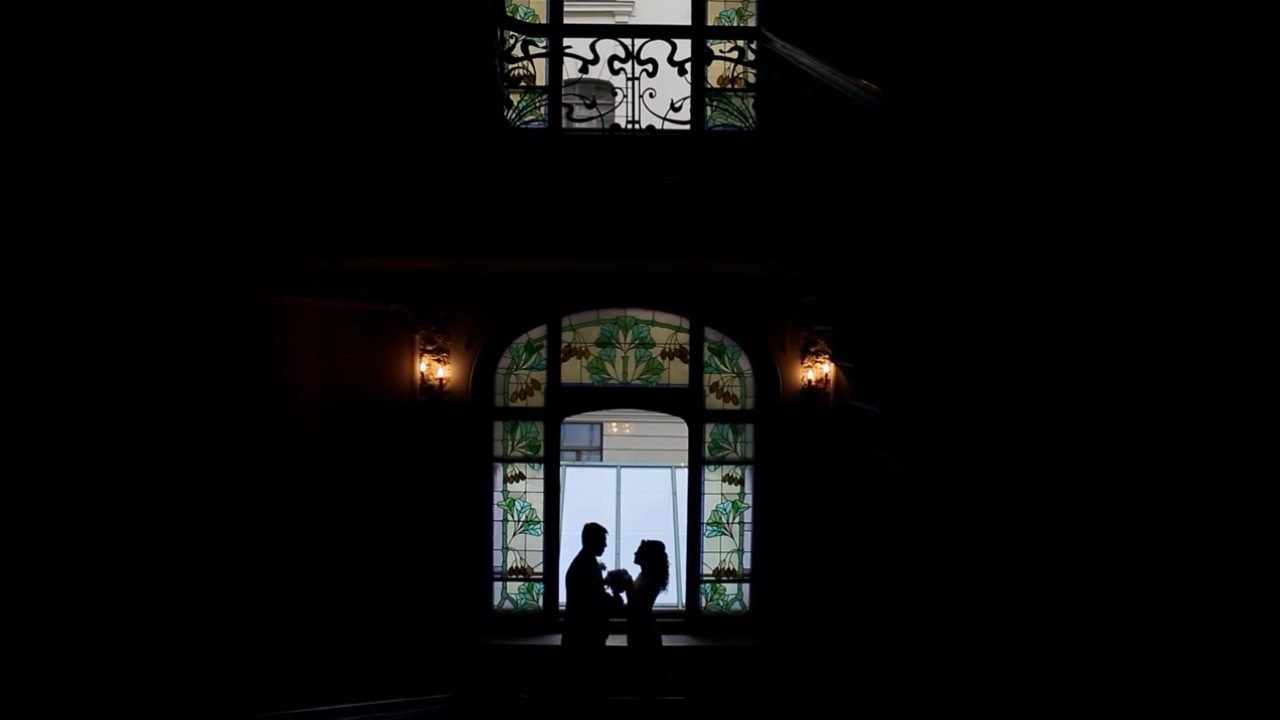 A young man and a young woman, a bride and groom, hold hands and smile while standing together indoors with a crowd of people around them.