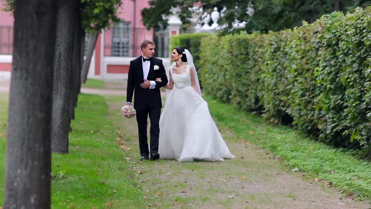 A bride and groom walk together and laugh as they hold hands on a tree-lined path.