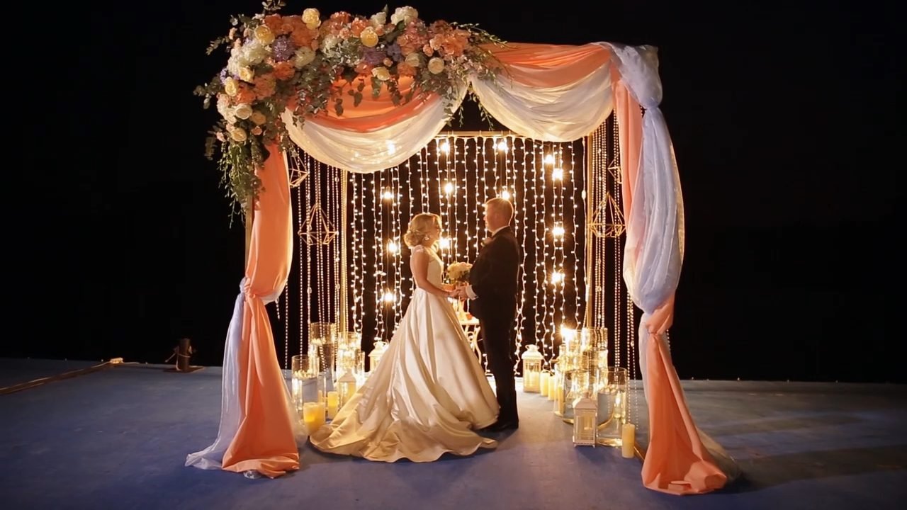 A bride, seen from the back, stands at the top of a grand, decorative stone staircase, holding a bouquet of pink flowers.