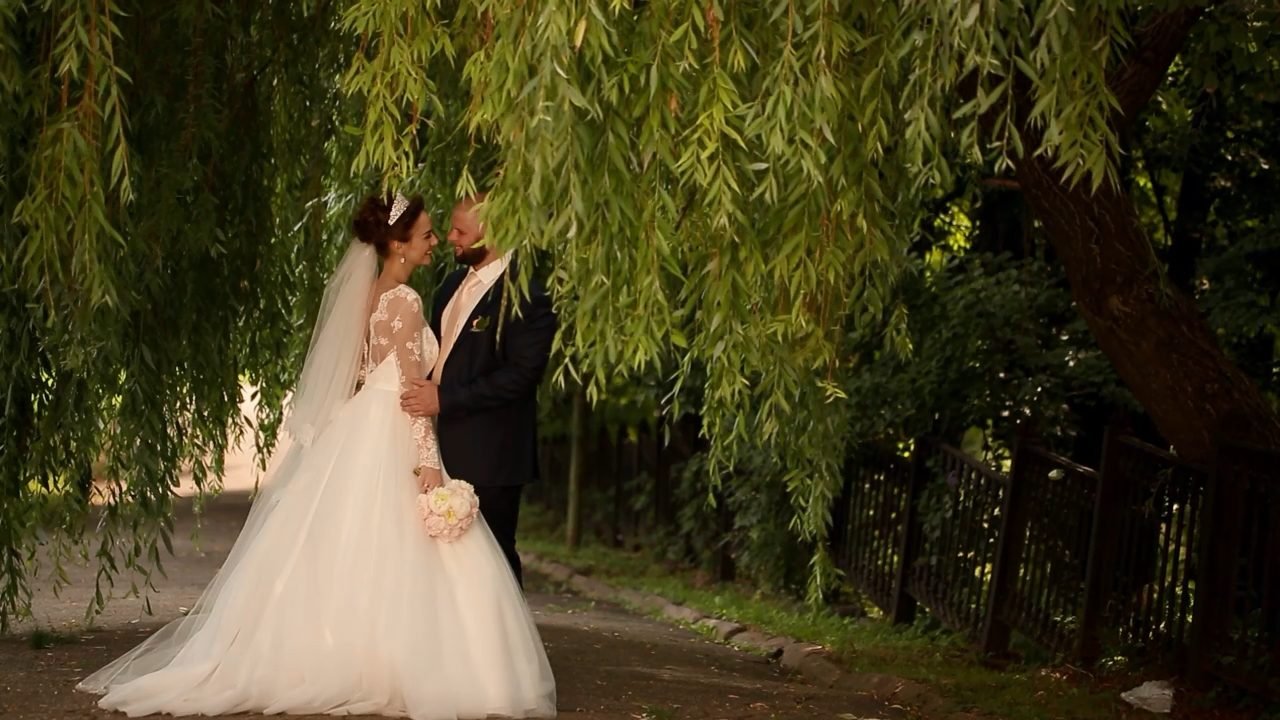 A bride and groom, Nancy and John, share a tender moment under the hanging branches of a willow tree. Nancy wears a white lace gown and tiara, holding a pink bouquet, while John is in a dark suit.